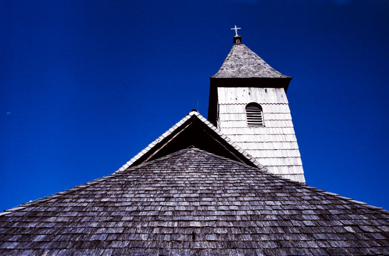 Close-up of a wooden church's steep shingle roof and bell tower against a clear blue sky in Austria.